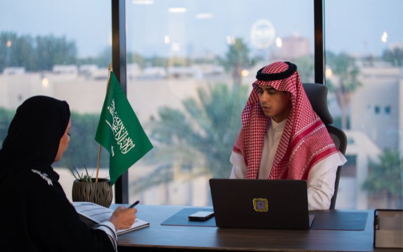 Saudi official in traditional attire sitting at a desk with a laptop, discussing with a woman wearing an abaya, with a Saudi flag on the table and a cityscape in the background